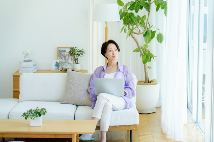 大きな窓のそばでソファに座ってひざの上にPCを広げる女性。