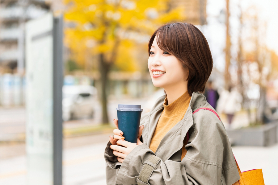 コーヒーを手に笑顔で外を歩く女性。背景には紅葉した街路樹。